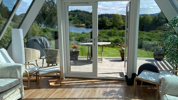 A conservatory with a wooden floor, white furniture, and a view of a lake and greenery.