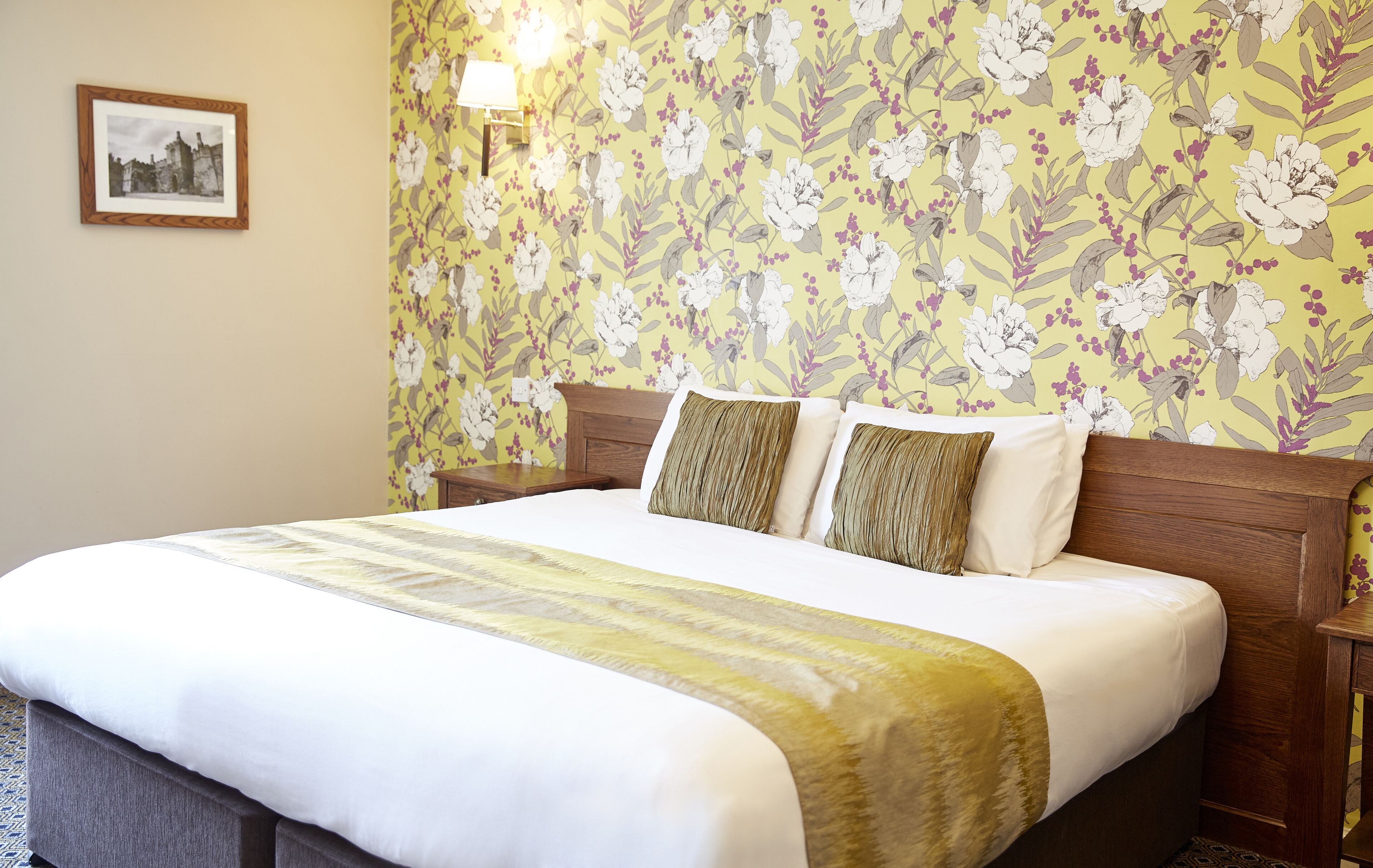 Una cama bien hecha con papel tapiz floral, un cuadro en la pared y una lámpara de noche.