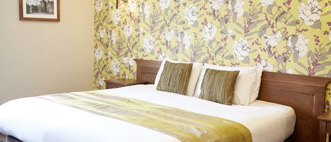 Una cama bien hecha con papel tapiz floral, un cuadro en la pared y una lámpara de noche.