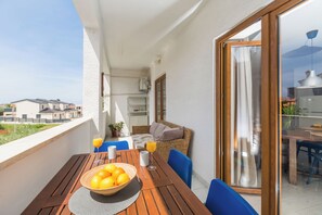 Un balcon avec une table en bois, des chaises bleues, un canapé en osier et une porte vitrée donnant sur un espace de repas intérieur.