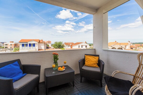 Un balcon agrémenté de meubles en osier, une petite table avec un vase rempli de fleurs et d’oranges, et une vue sur des bâtiments situés au loin.