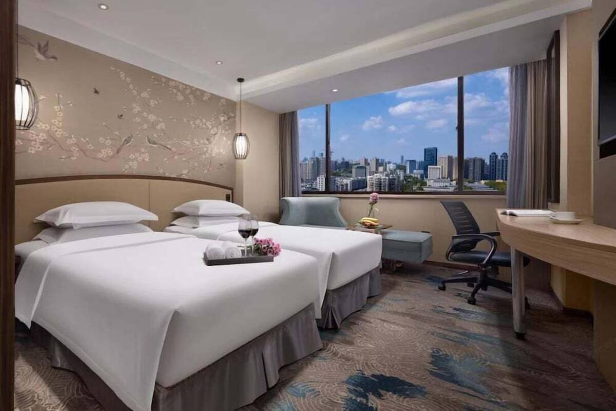 A hotel room with two beds, a desk, a chair, a large window with a city view, and a decorative wall.