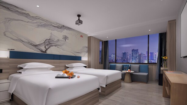 Habitación de hotel moderna con dos camas, vista a la ciudad, un amplio mural abstracto y un pequeño rincón de descanso.