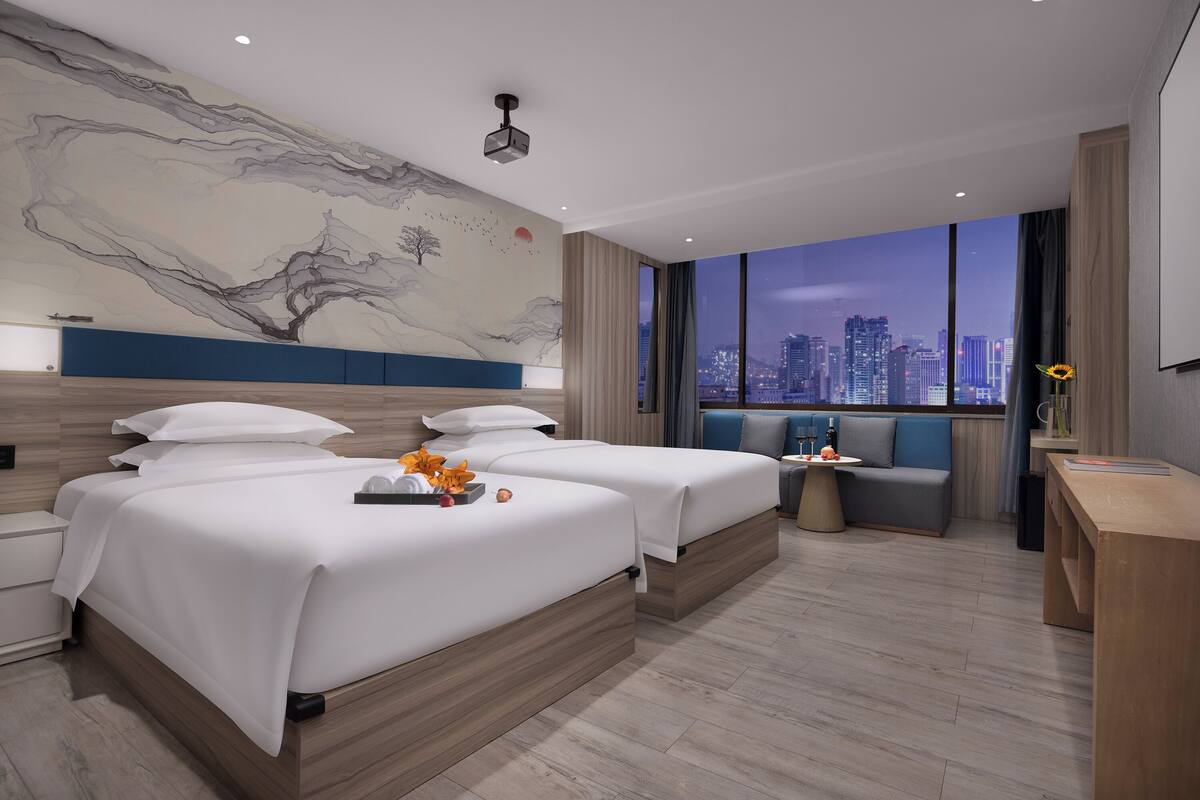 A modern hotel room with two beds, a city view, a large abstract mural, and a small seating area.