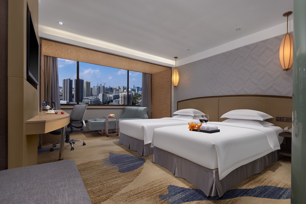 A hotel room with two beds, a desk, a chair, a TV, and a large window with a city view.
