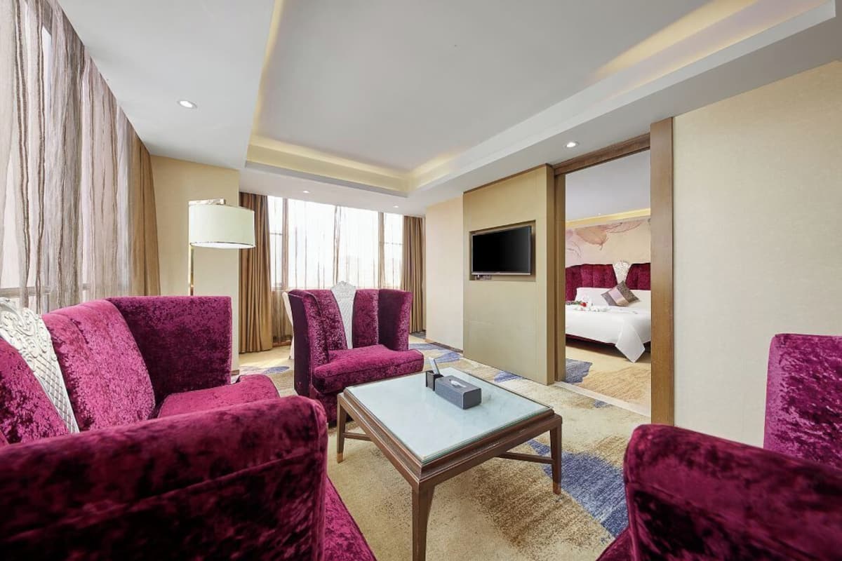 A hotel room with a large bed, a TV, a coffee table, and a seating area with red velvet chairs.