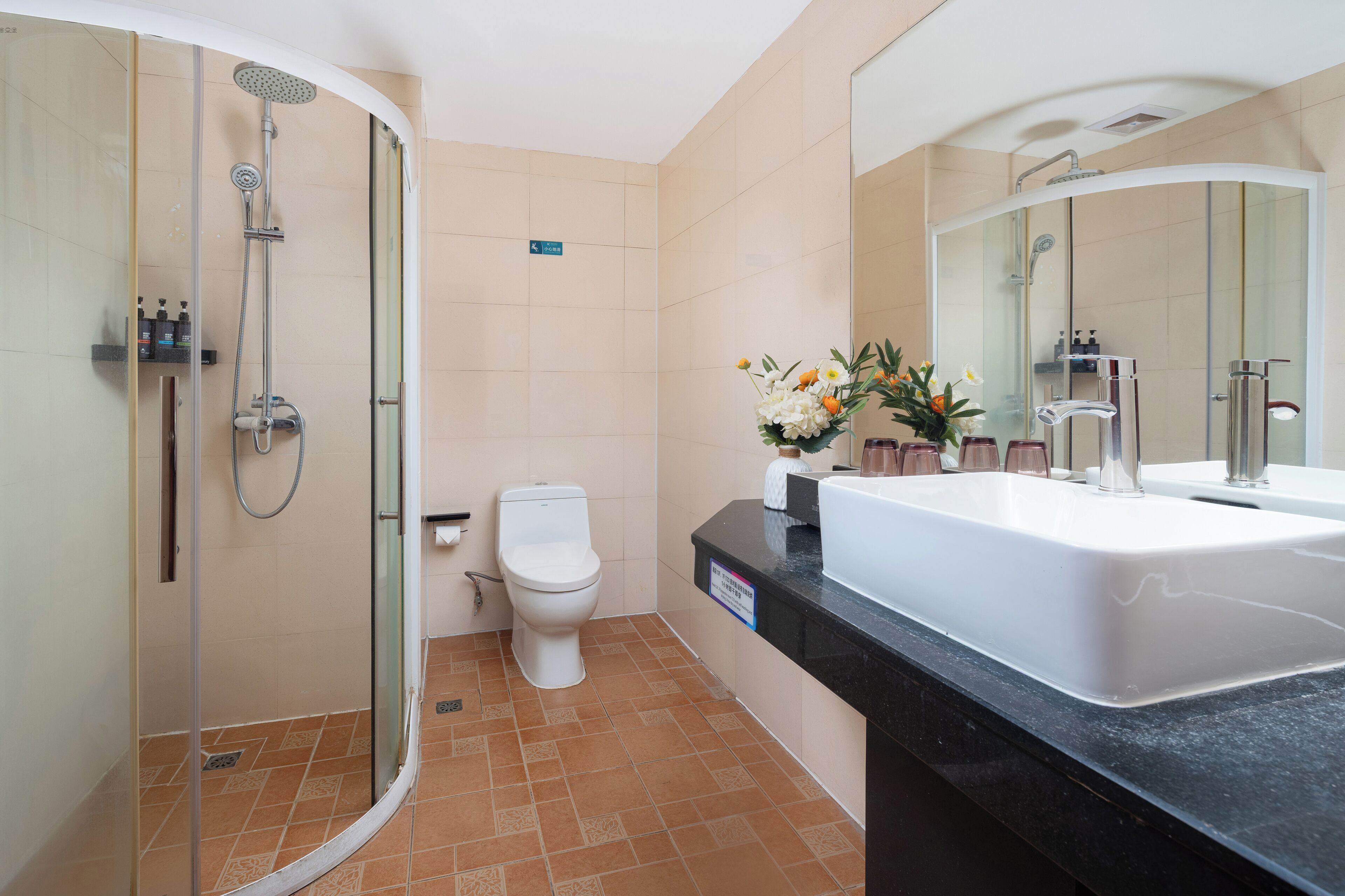 Un baño con ducha envuelta en vidrio, inodoro blanco, lavabo con espejo y un florero con flores sobre la encimera.