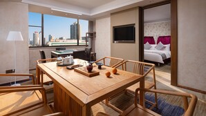 Habitación de hotel con comedor, cama con cortinas rosas, televisor y vista a la ciudad.