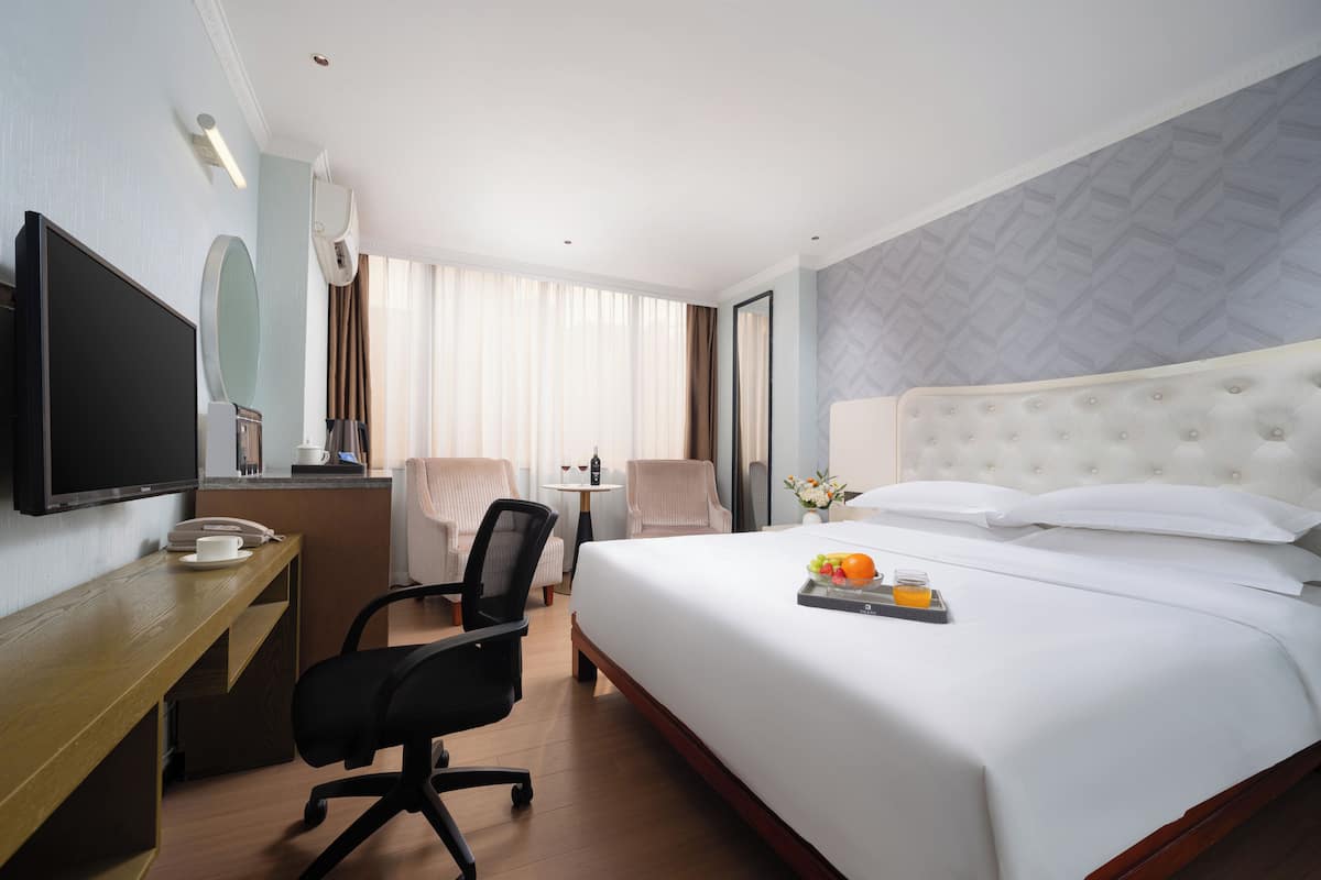 A hotel room with a large bed, a desk with a computer, a TV, and a small table with a cup and a phone.
