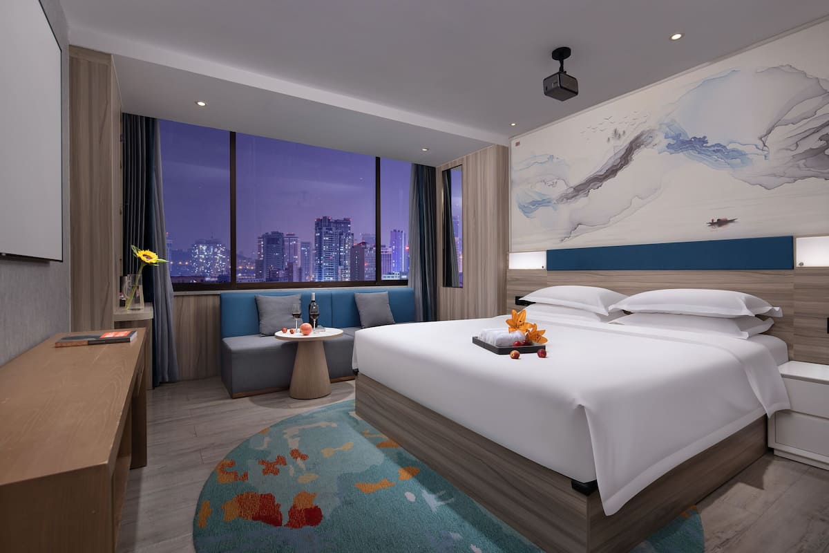 A modern hotel room with a large bed, a seating area, a small table, and a city view.