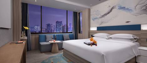 A modern hotel room with a large bed, a seating area, a small table, and a city view.