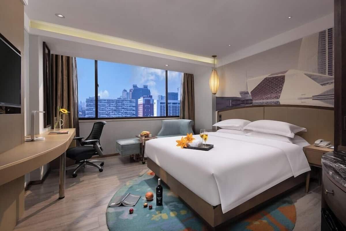 A hotel room with a large bed, a desk, a chair, a small table, a nightstand, a lamp, and a view of the cityscape.