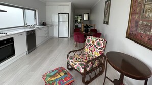 Moderni keittiö, jossa on valkoiset kaapit, sisäänrakennettu astianpesukone ja jääkaappi. Ruokailutilassa on puinen pöytä ja punaiset tuolit. Värikäs tuoli ja rahi on sijoitettu kuvan etualaan.