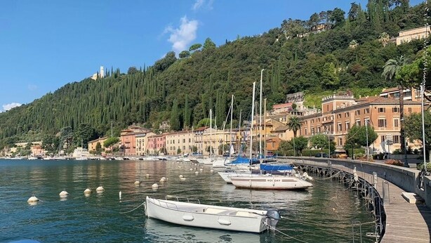 A coastal town with boats docked along a wooden pier, buildings with balconies overlooking the water, and a hill with dense greenery in the background. - Old town Villa Rustico vacation home 600 meters to the SEE CASA LINDNER 14Guest (Maderno)