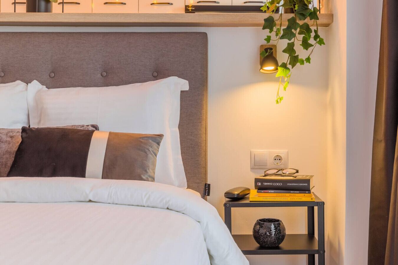 Un lit bien fait, avec une literie blanche et marron, une table de chevet avec une lampe, un vase et une pile de livres.