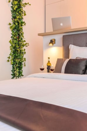 A neatly made bed with a laptop on a wooden shelf above it, a bottle of wine, and glasses on the nightstand.