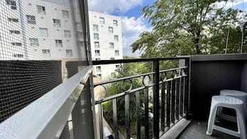 Ein Balkon mit einem Maschendrahtgeländer, einem kleinen weißen Hocker und Blick auf Gebäude und Bäume.