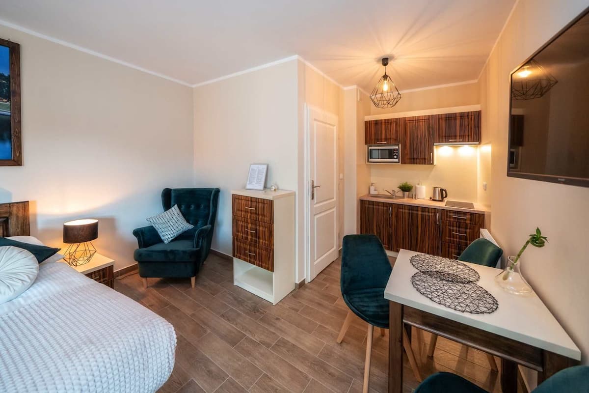 A compact hotel room with a bed, a chair, a small table, a kitchenette, and a mirror.