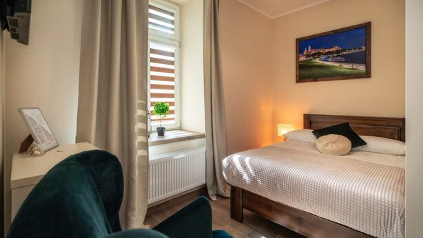 A hotel room with a bed, a window with blinds, a potted plant, a painting on the wall, and a desk with a sign. - Apartments 17 Szewska (Kraków)
