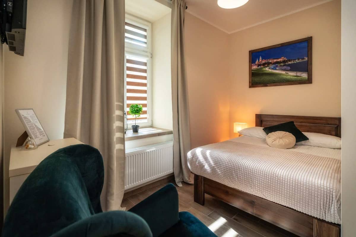 A hotel room with a bed, a window with blinds, a potted plant, a painting on the wall, and a desk with a sign.