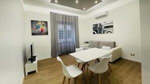 A modern living room with a dining area, featuring a white table and chairs, a grey sofa, and a large window with curtains. - Home Vatican Oasis (Roma)