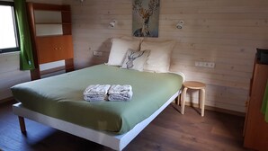 Un lit simple avec une couvre-lit verte, une tête de lit en bois, un petit tabouret et un tableau représentant un cerf accroché au mur.