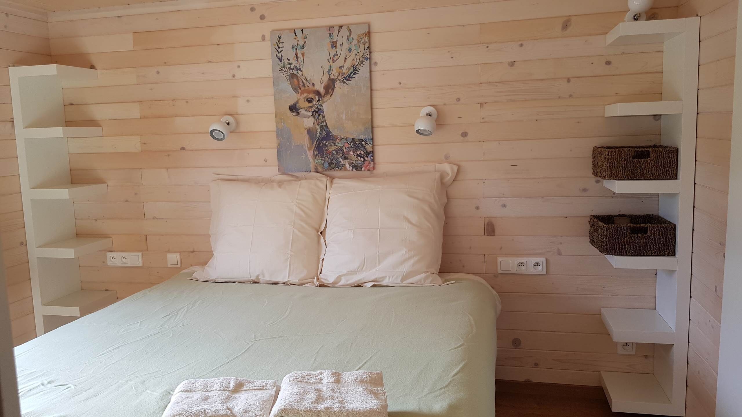 A bedroom with a bed, two pillows, a painting, wall-mounted lights, and storage shelves.