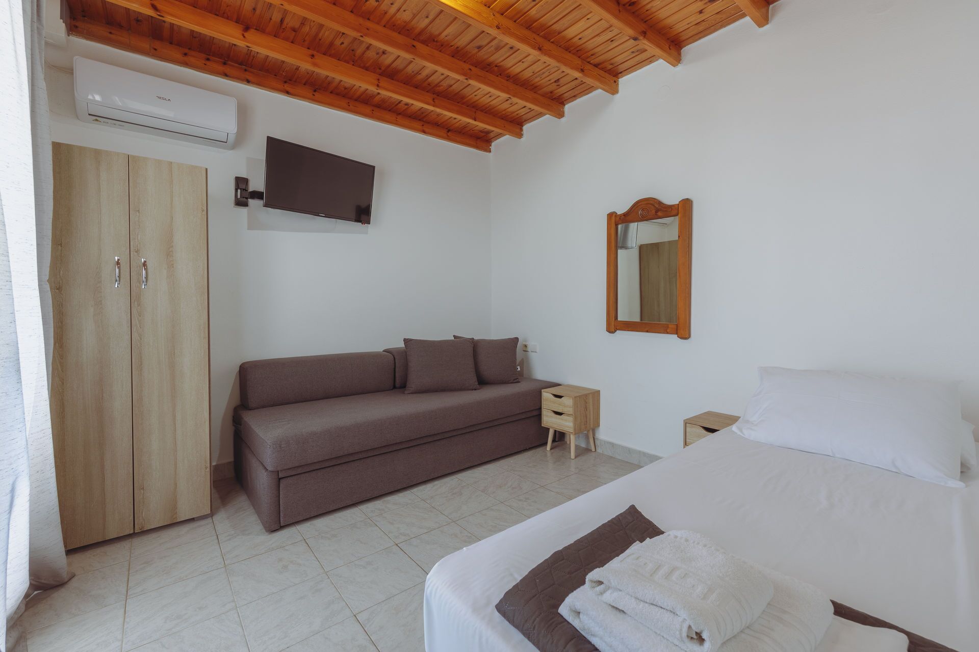 Een slaapkamer met een bed, een bank, een houten garderobe, een nachtkastje, een spiegel en een aan de muur bevestigde televisie.