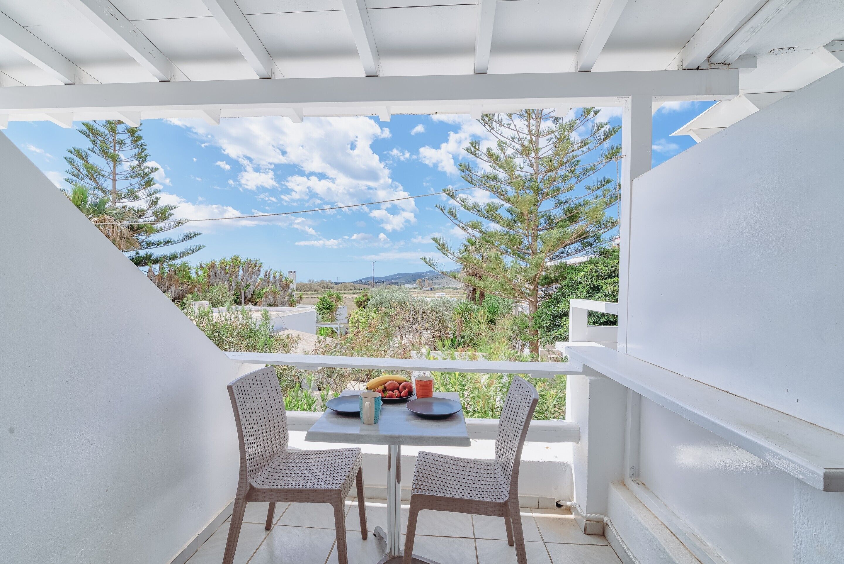 A balcony with a white railing, two wicker chairs, a small table, and a view of trees and distant hills.