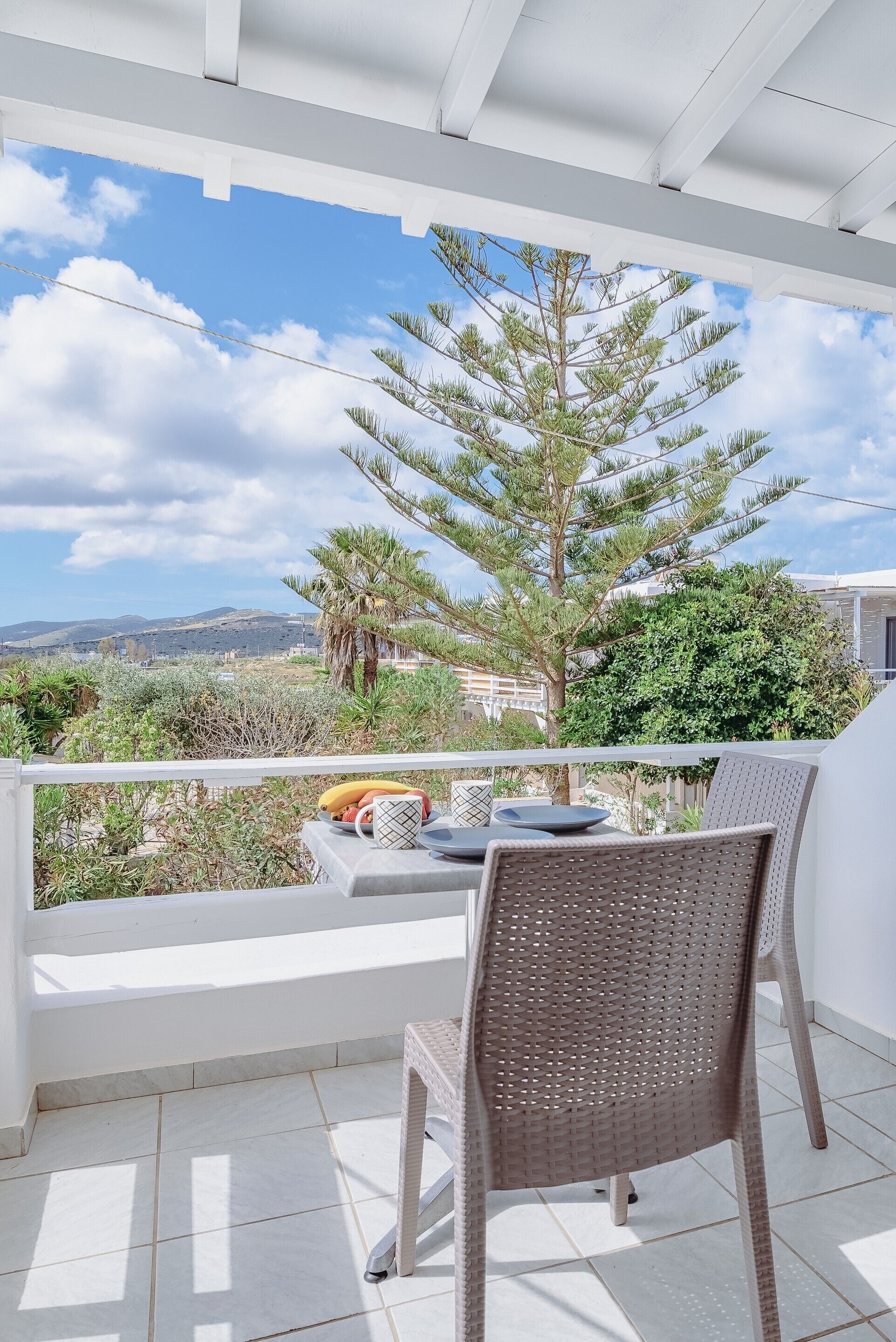 A balcony with a wicker chair and a table, overlooking a landscape with trees and distant hills.