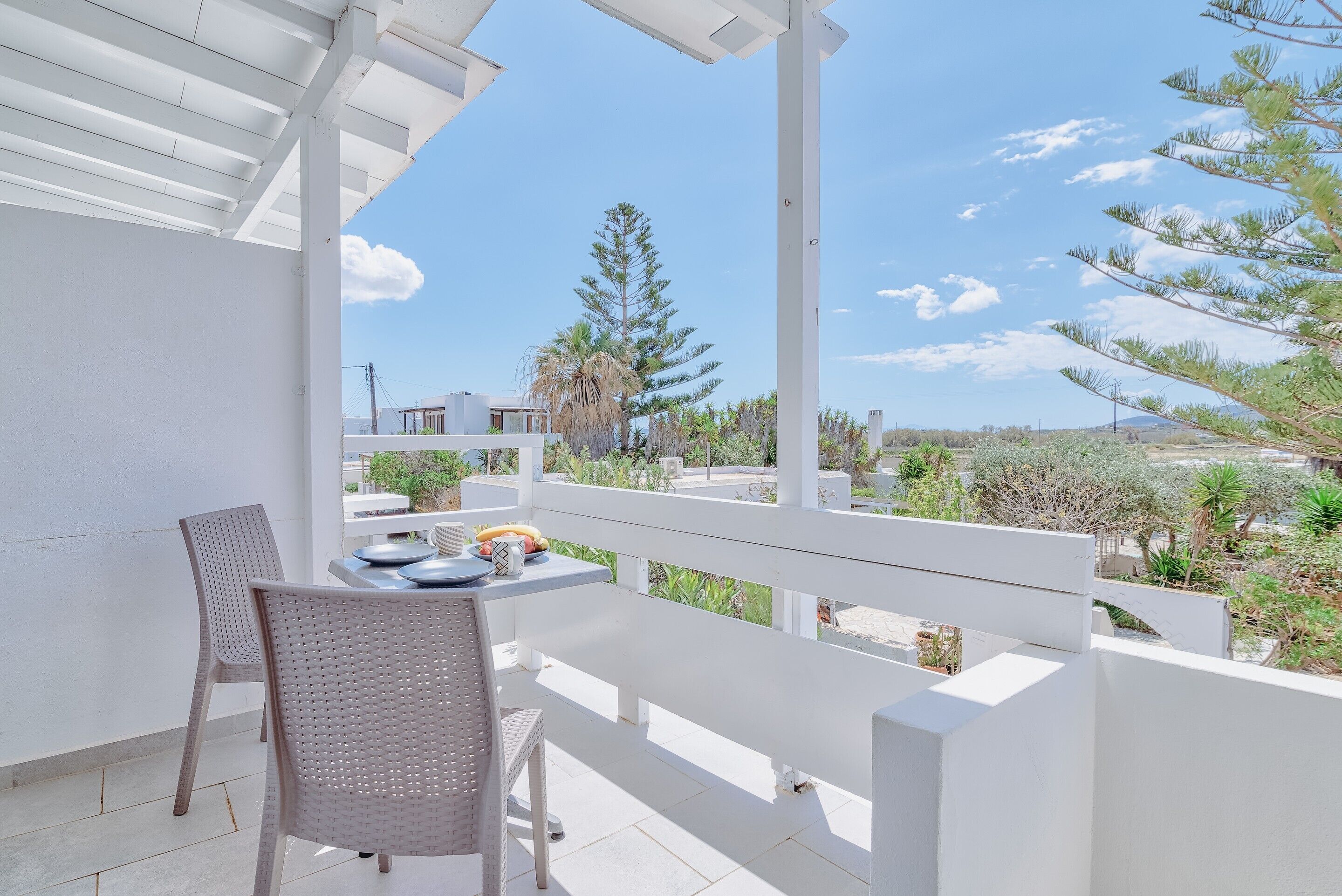 A balcony with a white table and chairs, overlooking a garden with trees and a clear sky.