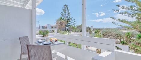 A balcony with a white table and chairs, overlooking a garden with trees and a clear sky.