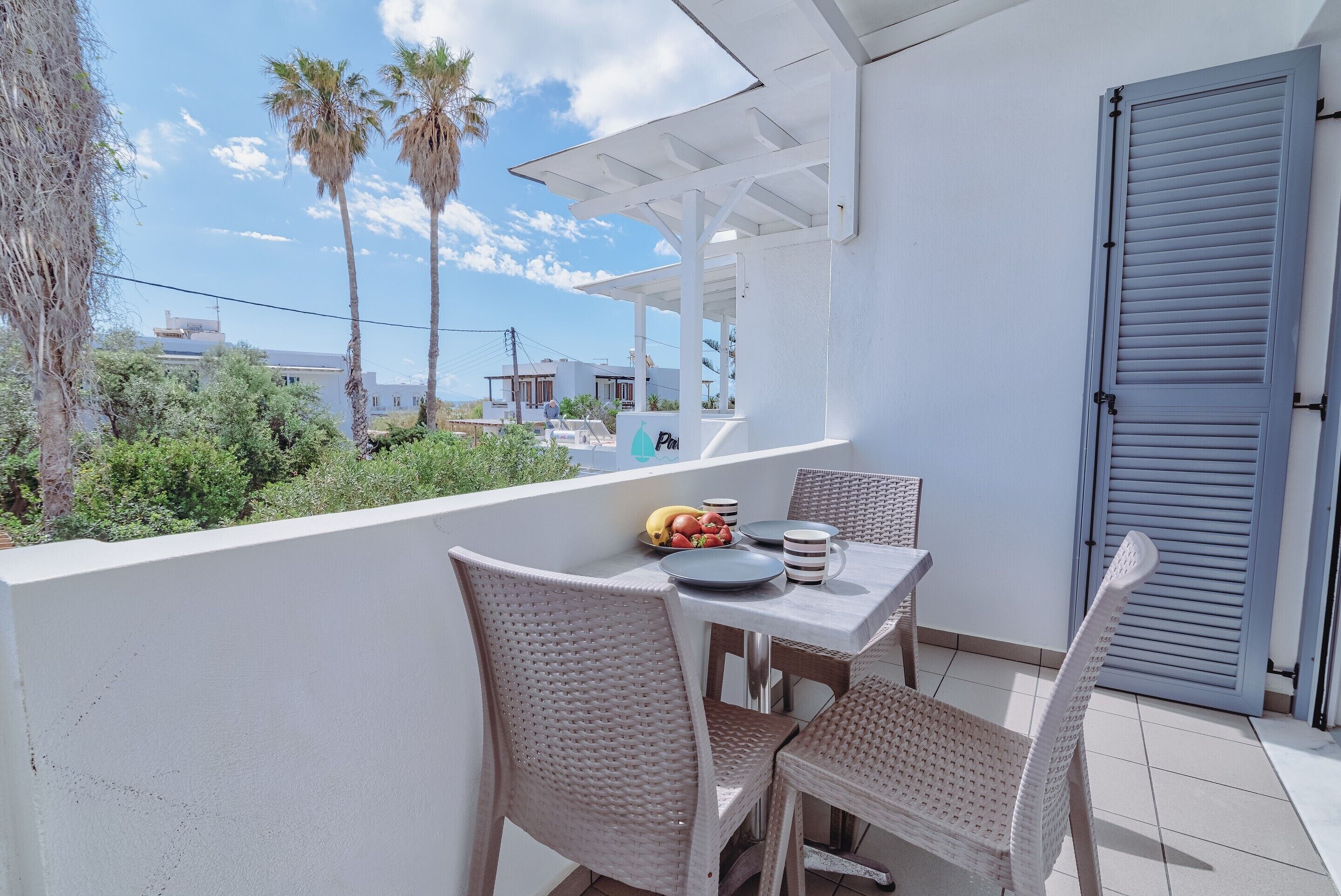 Un balcón con sillas de mimbre y una mesa dispuesta para dos, con vistas a una zona residencial con palmeras y casas.
