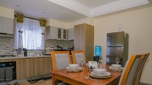 A dining area with a wooden table set for four, surrounded by chairs. A kitchen with modern appliances and a water dispenser is visible in the background. - Kijala Suite by Homes By Della (Nairobi)
