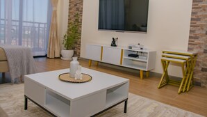 A modern living room with a white coffee table, a flat-screen TV mounted on the wall, a white cabinet with gold accents, and a yellow metal side table. - Kijala Suite by Homes By Della (Nairobi)