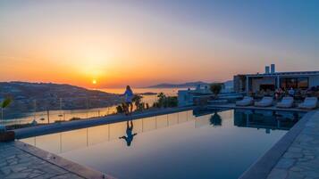 Una piscina infinita en la azotea con vistas a la puesta de sol sobre un cuerpo de agua, sillas de descanso y un edificio con grandes ventanales.
