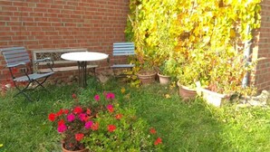 Um pátio com muro de tijolos, duas esplanadas azuis, uma mesa redonda e vasos com plantas de flores vermelhas.