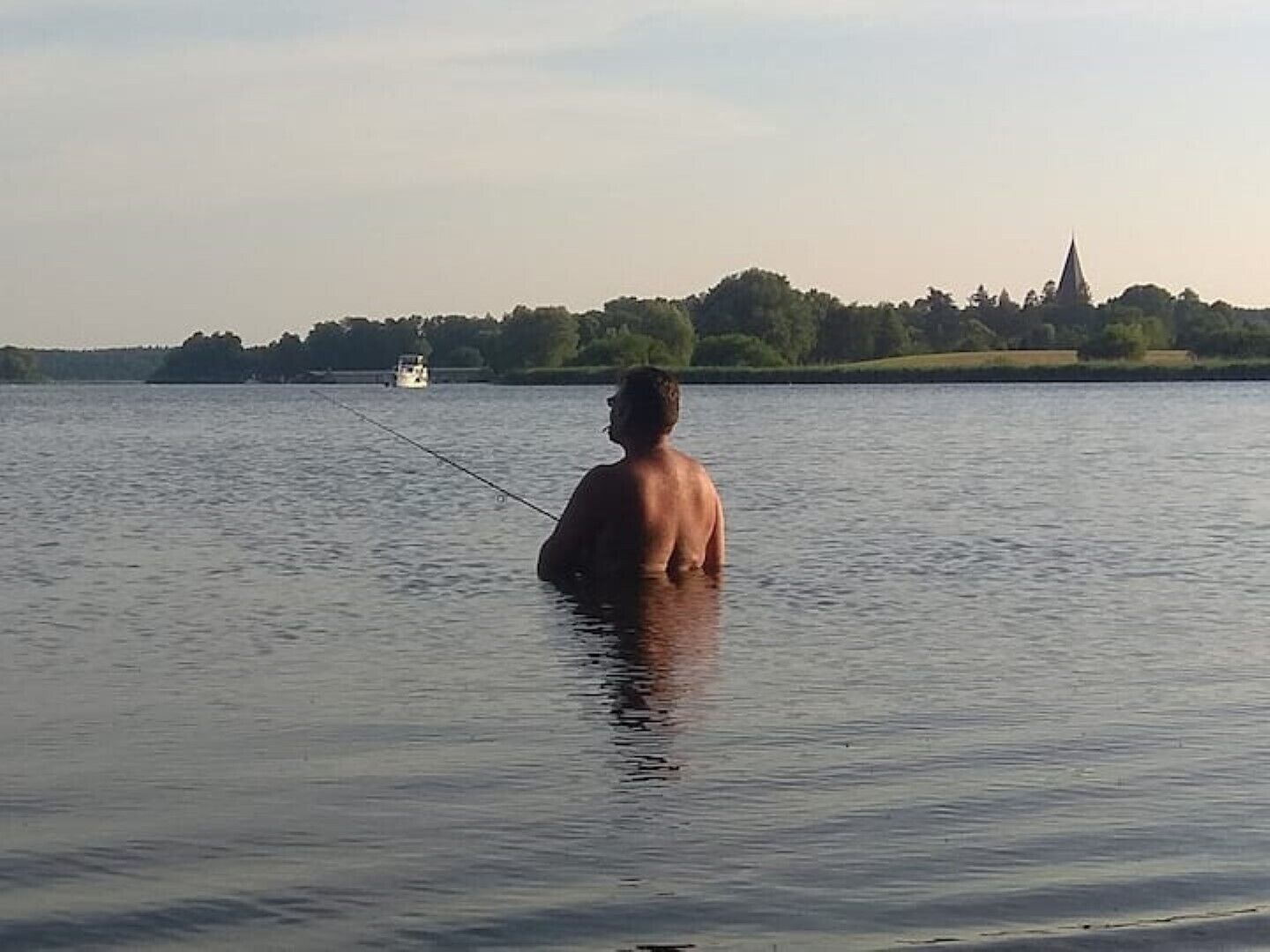 Ein Mann beim Angeln in einem ruhigen See, im Hintergrund ein Boot und am fernen Ufer eine Kirche.