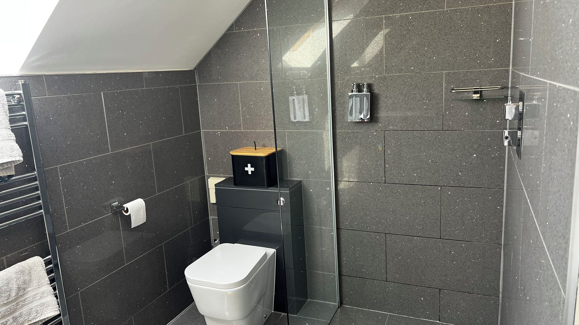 A modern bathroom with dark tiles, a white toilet, a black cabinet with a wooden top, and a glass shower enclosure.