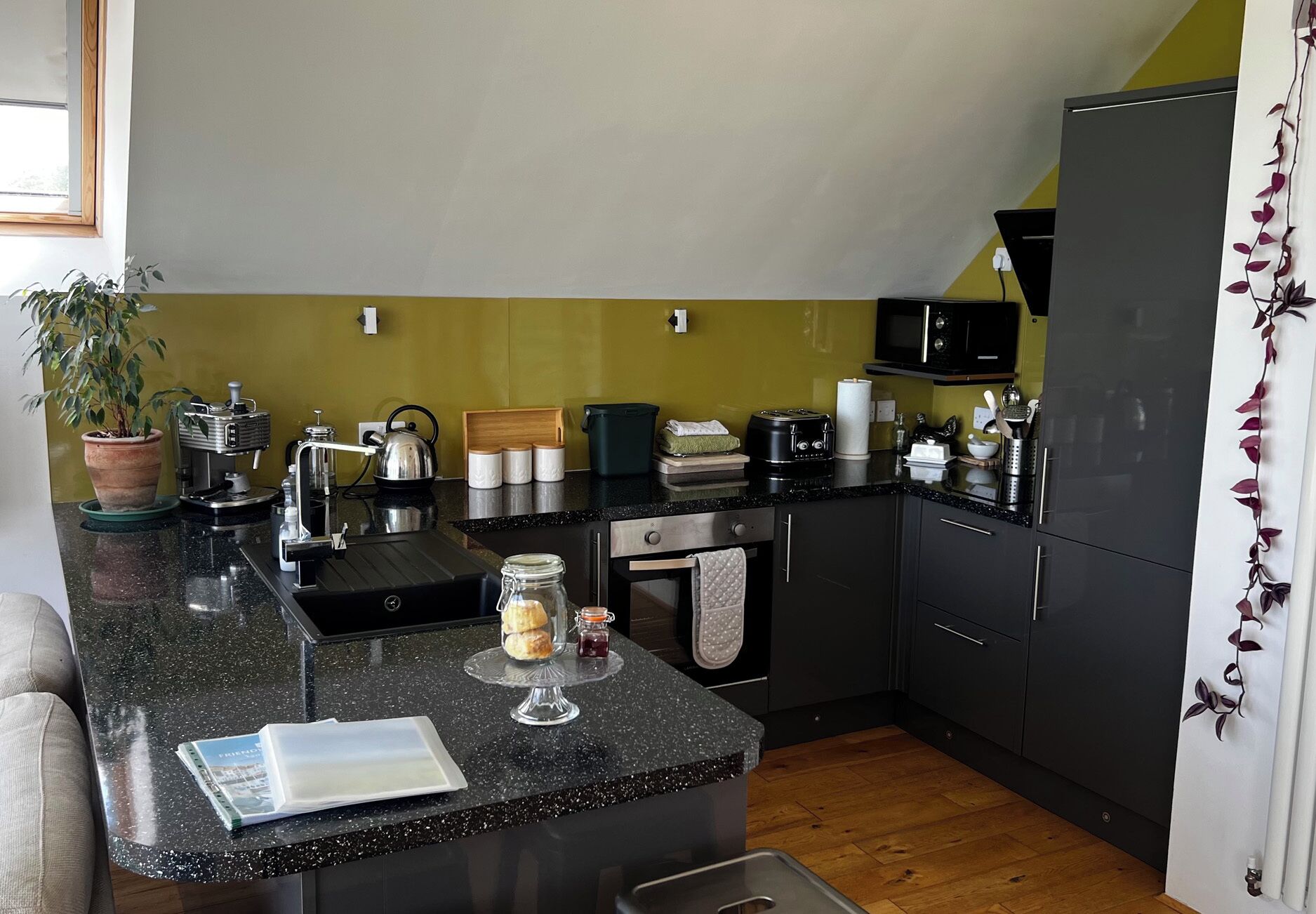 A compact kitchen with a coffee machine, toaster, kettle, and a small island with a glass jar and scones.