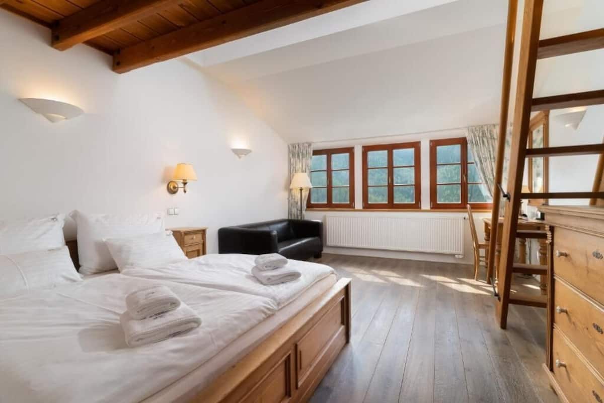 Quarto com cama de madeira, duas luminárias de cabeceira, um sofá preto, cômoda de madeira e janela com cortinas.