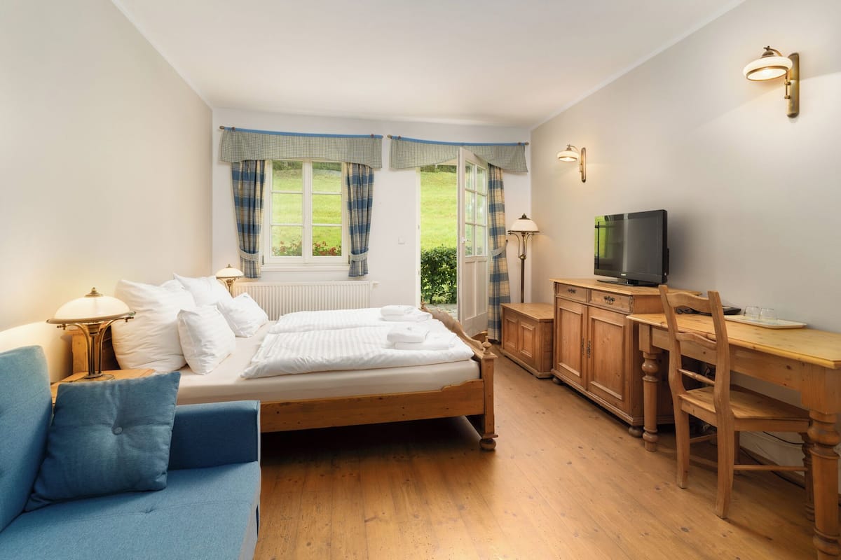 Quarto com cama, escrivaninha, cadeira, televisão e janela com cortinas.