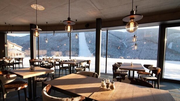 Un restaurant avec des tables et des chaises en bois, de grandes fenêtres donnant sur les montagnes et des lampes suspendues.