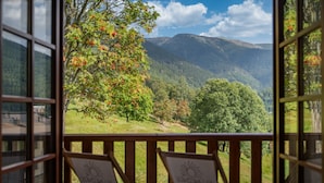 Un balcon doté de balustrades en bois et de deux fauteuils de jardin portant un logo, offrant une vue sur un paysage montagneux avec des arbres et un ciel dégagé.