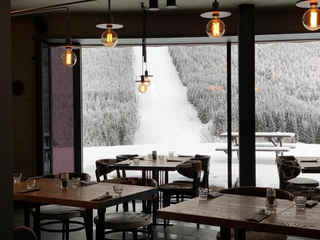 A restaurant with tables and chairs, large windows with a snowy view, and hanging pendant lights.