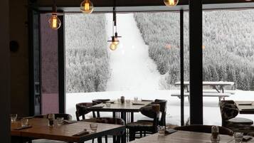 A restaurant with tables and chairs, large windows with a snowy view, and hanging pendant lights.