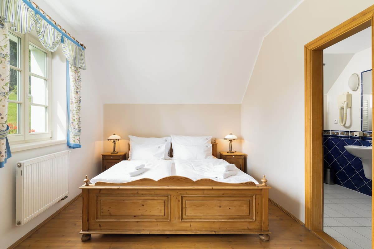 Quarto com cama de madeira, duas luminárias de cabeceira, janela com cortinas florais e vista para o banheiro por uma porta aberta.