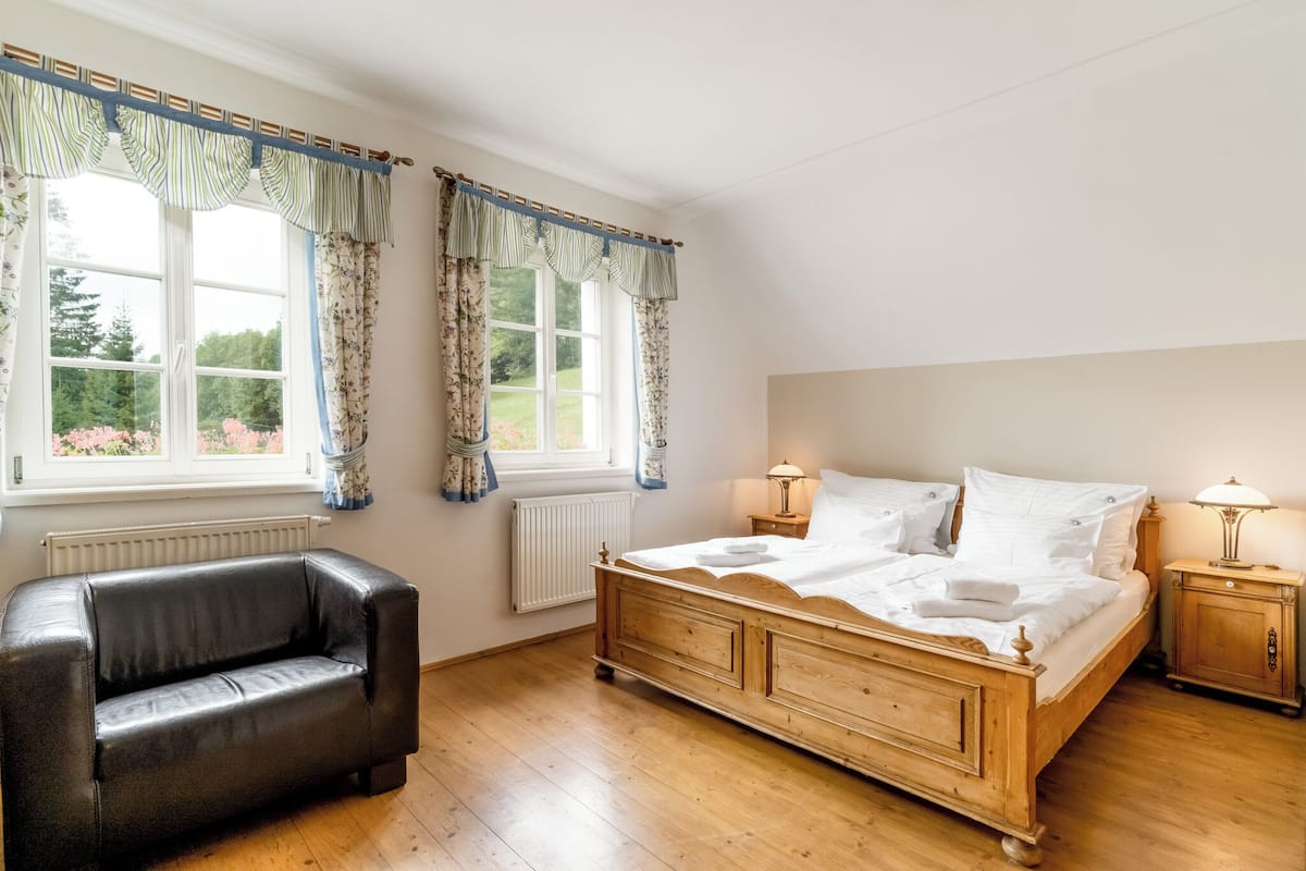 Quarto com cama de madeira, duas mesas de cabeceira com luminárias, uma poltrona de couro preto e uma janela com cortinas.