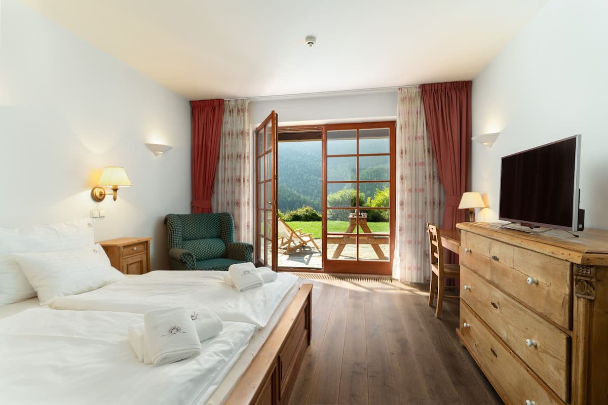 Quarto de hotel com cama, televisão, cómoda de madeira, cadeira e vista para um jardim através de uma porta aberta.