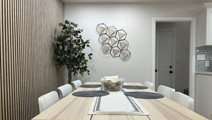 Ein Essbereich mit einem Holztisch, weißen Stühlen, einem Wandkunstwerk und einer Topfpflanze.